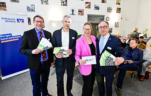 Vier Personen bei einer Veranstaltung in einem Raum, lächelnd und Broschüren haltend.