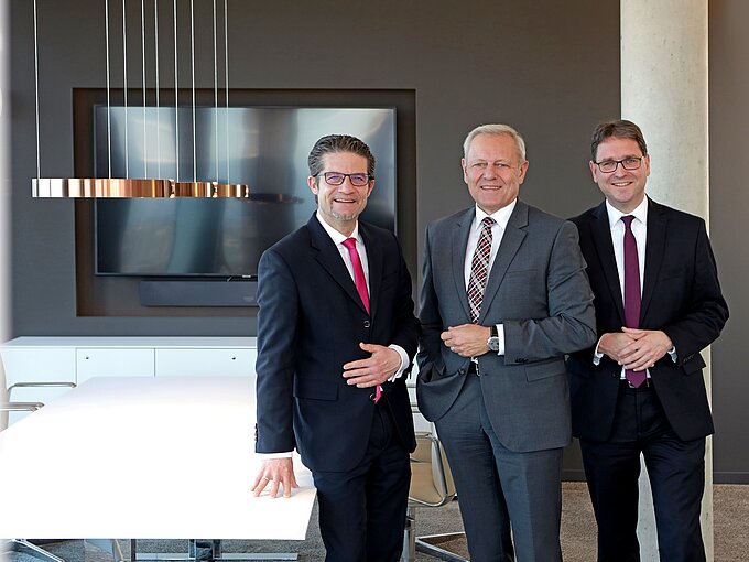 Drei Männer in Anzügen stehen lächelnd in einem modernen Konferenzraum mit Tisch und Bildschirm im Hintergrund.