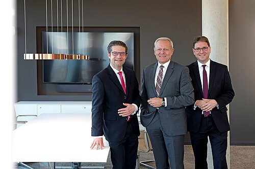 Drei Männer in Anzügen stehen lächelnd in einem modernen Konferenzraum mit Tisch und Bildschirm im Hintergrund.
