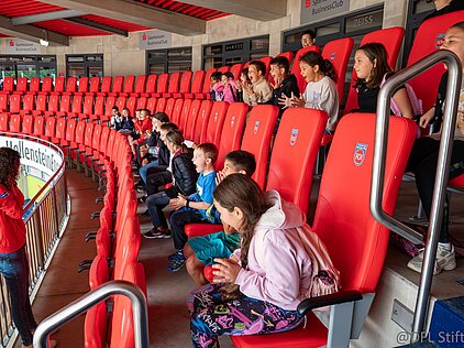 Kinder sitzen in roten Stadionstühlen, hören aufmerksam einer Frau zu.
