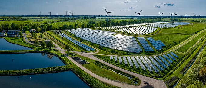 Luftaufnahme eines großen Solarparks mit Windturbinen im Hintergrund, umgeben von grüner Landschaft und Teichen.