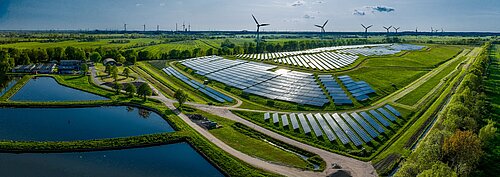 Luftaufnahme eines großen Solarparks mit Windturbinen im Hintergrund, umgeben von grüner Landschaft und Teichen.