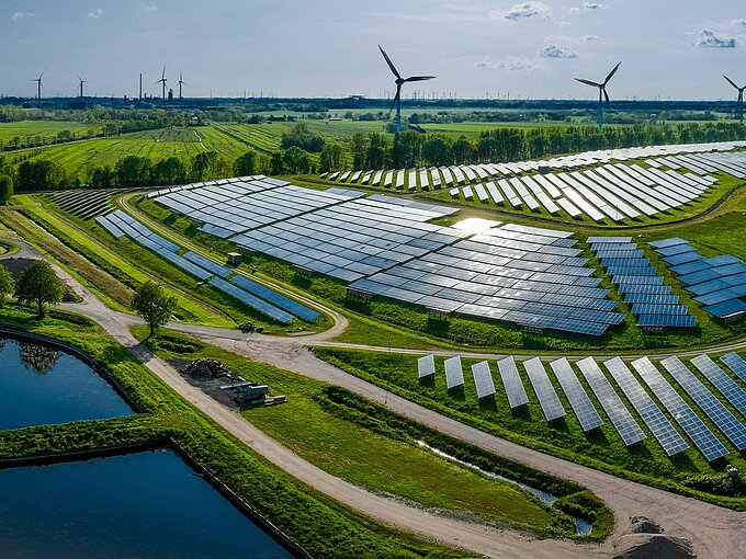 Luftaufnahme eines großen Solarparks mit Windturbinen im Hintergrund, umgeben von grüner Landschaft und Teichen.