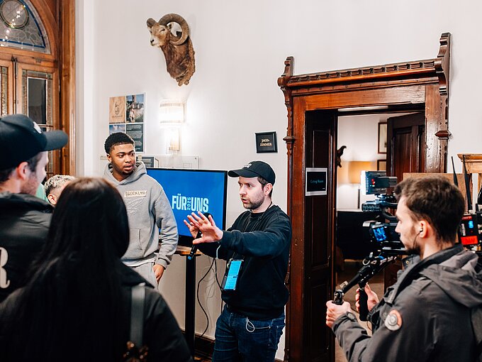 Filmcrew bei der Arbeit in einem Raum mit Jagdtrophäe an der Wand und einem Bildschirm, der "Für Uns" zeigt.