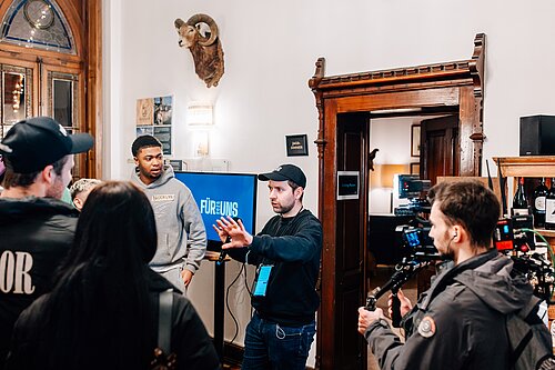 Filmcrew bei der Arbeit in einem Raum mit Jagdtrophäe an der Wand und einem Bildschirm, der "Für Uns" zeigt.