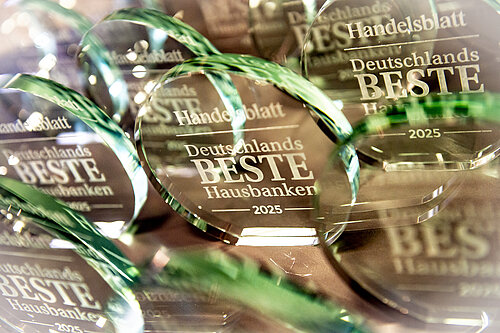 Das Bild zeigt mehrere runde Glas-Awards mit der Gravur "Handelsblatt Deutschlands beste Hausbanken 2025". Die Awards sind aus klarem Glas und reflektieren das Licht, wodurch ein schimmernder Effekt entsteht. Sie liegen dicht beieinander auf einer Oberfläche.