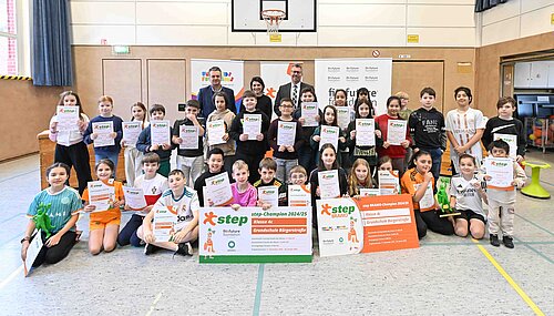 Kindergartenkinder halten Urkunden und große Plakate in einer Turnhalle, umgeben von Lehrern.