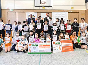 Kindergartenkinder halten Urkunden und große Plakate in einer Turnhalle, umgeben von Lehrern.