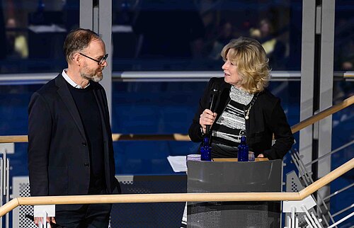 Zwei Personen stehen auf einer Bühne. Eine Frau spricht in ein Mikrofon und lächelt, ein Mann hört aufmerksam zu.