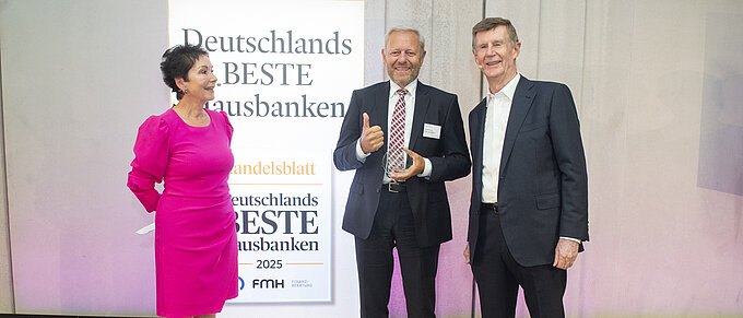 Drei Personen stehen vor einem Schild mit der Aufschrift "Deutschlands Beste Hausbanken 2025". Die Person links trägt ein pinkes Kleid und hat die Hände hinter dem Rücken. Die Person in der Mitte hält eine Auszeichnung und zeigt den Daumen hoch, während sie einen dunklen Anzug mit weißem Hemd und rot karierter Krawatte trägt. Die Person rechts hat einen dunklen Anzug und weißes Hemd an. Alle drei lächeln in die Kamera.