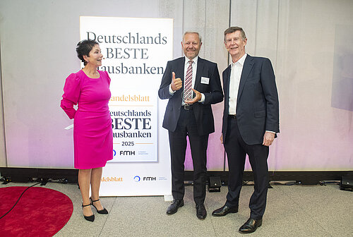 Drei Personen stehen vor einem Schild mit der Aufschrift "Deutschlands Beste Hausbanken 2025". Die Person links trägt ein pinkes Kleid und hat die Hände hinter dem Rücken. Die Person in der Mitte hält eine Auszeichnung und zeigt den Daumen hoch, während sie einen dunklen Anzug mit weißem Hemd und rot karierter Krawatte trägt. Die Person rechts hat einen dunklen Anzug und weißes Hemd an. Alle drei lächeln in die Kamera.
