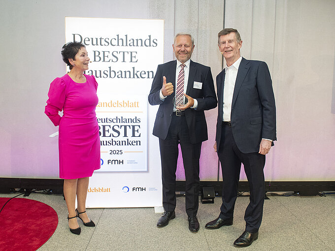 Drei Personen stehen vor einem Schild mit der Aufschrift "Deutschlands Beste Hausbanken 2025". Die Person links trägt ein pinkes Kleid und hat die Hände hinter dem Rücken. Die Person in der Mitte hält eine Auszeichnung und zeigt den Daumen hoch, während sie einen dunklen Anzug mit weißem Hemd und rot karierter Krawatte trägt. Die Person rechts hat einen dunklen Anzug und weißes Hemd an. Alle drei lächeln in die Kamera.