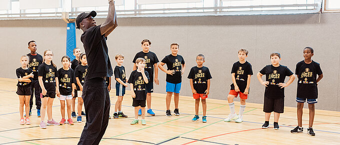 Dennis Schröder wirft einen Basketball, während Kinder in schwarz-gelben Shirts in einer Turnhalle zuschauen.