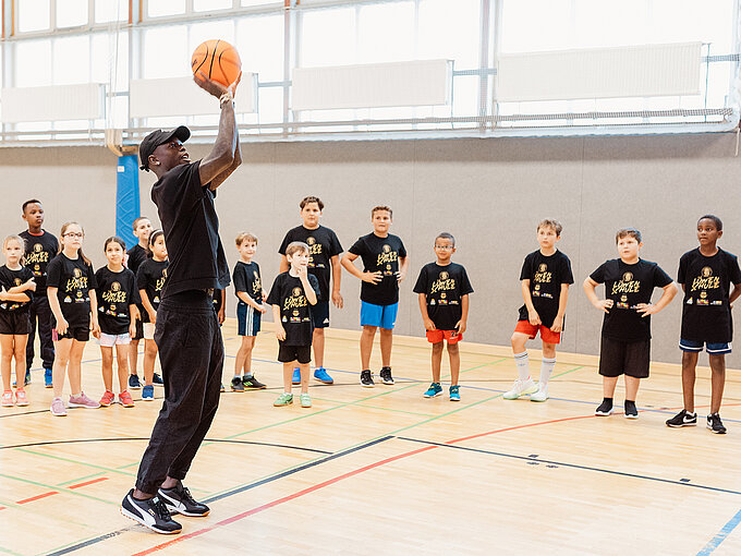 Basketball-Training für Kinder in der Sporthalle Dennis Schröder wirft einen Basketball, während Kinder in schwarz-gelben Shirts in einer Turnhalle zuschauen.