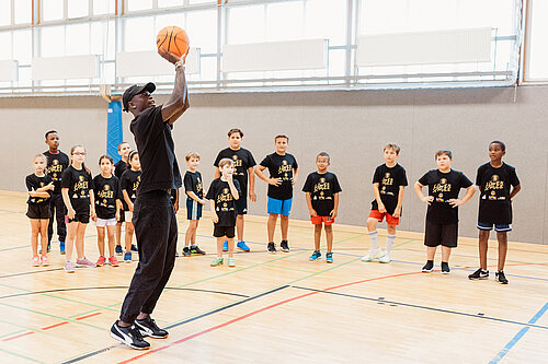 Basketball-Training für Kinder in der Sporthalle Dennis Schröder wirft einen Basketball, während Kinder in schwarz-gelben Shirts in einer Turnhalle zuschauen.