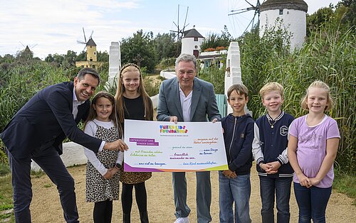 Eine Gruppe von sechs Kindern und zwei Erwachsenen steht draußen in einem parkähnlichen Bereich mit Windmühlen im Hintergrund. Die Erwachsenen halten ein großes Plakat mit der Aufschrift "unterstützt uns Kinder dabei..." und mehreren farbenfrohen Satzfragmenten. Die Kinder lächeln und stehen in einer Reihe, während die Erwachsenen sich zu ihnen lehnen. Die Szene wirkt fröhlich und positiv.