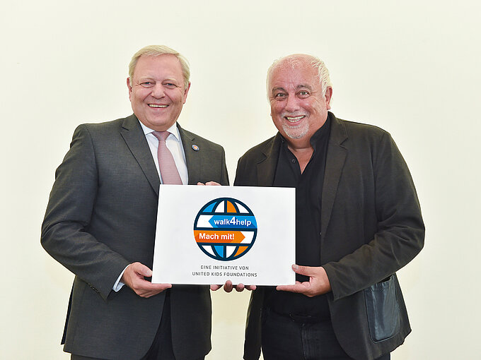 Zwei Männer stehen nebeneinander vor einem neutralen, hellen Hintergrund und halten gemeinsam ein Schild mit dem Logo und Slogan der Initiative "walk4help" in den Händen. Der Mann auf der linken Seite trägt einen dunklen Anzug mit weißem Hemd und einer hellrosa Krawatte. Er hat kurzes, blond-graues Haar und lächelt freundlich. Der Mann auf der rechten Seite trägt eine schwarze Jacke über einem schwarzen Hemd, hat kurze, weiße Haare und ein Lächeln im Gesicht. Das Schild zeigt eine stilisierte Weltkugel mit den Worten "walk4help" in der Mitte und "Mach mit!" darunter. Darunter steht der Hinweis, dass es sich um eine Initiative der United Kids Foundations handelt.