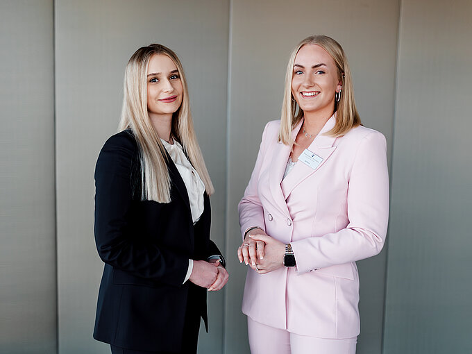 Auf dem Foto sieht man zwei junge blonde Frauen, die in die Kamera lächeln. Die linke Frau trägt einen dunklen Hosenanzug und ein weißes Hemd. Die rechte Frau trägt einen rosafarbenden Hosenanzug.