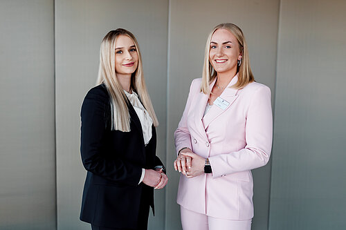 Auf dem Foto sieht man zwei junge blonde Frauen, die in die Kamera lächeln. Die linke Frau trägt einen dunklen Hosenanzug und ein weißes Hemd. Die rechte Frau trägt einen rosafarbenden Hosenanzug.