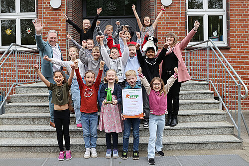 Eine fröhliche Kindergruppe steht mit zwei Erwachsenen jubelnd auf einer Treppe vor einem Schulgebäude. Ein Junge in der ersten Reihe hält stolz eine Urkunde, ein anderes Kind eine grüne Trophäe. Alle heben die Arme in die Luft und lachen, offensichtlich anlässlich einer Siegerehrung oder eines besonderen Erfolges.