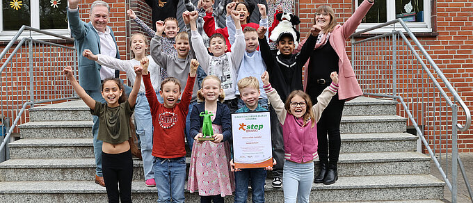Eine fröhliche Kindergruppe steht mit zwei Erwachsenen jubelnd auf einer Treppe vor einem Schulgebäude. Ein Junge in der ersten Reihe hält stolz eine Urkunde, ein anderes Kind eine grüne Trophäe. Alle heben die Arme in die Luft und lachen, offensichtlich anlässlich einer Siegerehrung oder eines besonderen Erfolges.
