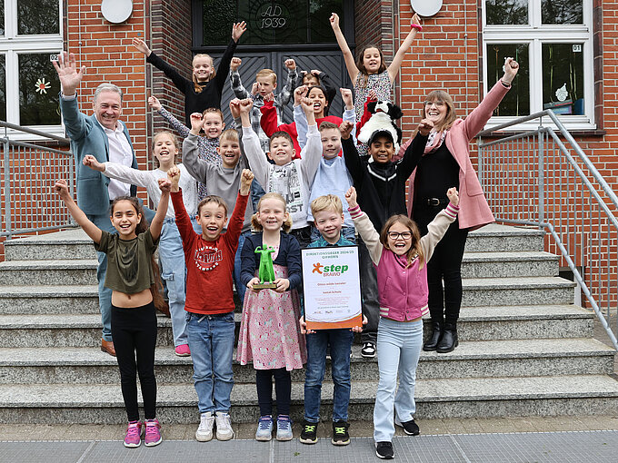 Eine fröhliche Kindergruppe steht mit zwei Erwachsenen jubelnd auf einer Treppe vor einem Schulgebäude. Ein Junge in der ersten Reihe hält stolz eine Urkunde, ein anderes Kind eine grüne Trophäe. Alle heben die Arme in die Luft und lachen, offensichtlich anlässlich einer Siegerehrung oder eines besonderen Erfolges.