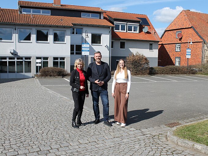 Drei Personen stehen lächelnd vor einem modernen Gebäude mit orangefarbenen Dächern an einem sonnigen Tag.
