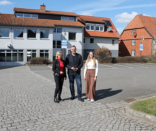 Drei Personen stehen lächelnd vor einem modernen Gebäude mit orangefarbenen Dächern an einem sonnigen Tag.