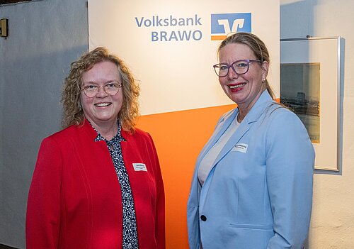 Auf dem Foto sieht man zwei Frauen, die in die Kamera lächeln. Die Frau links hat blonde lockige Haare, trägt eine Brille und ein rotes Jacket mit einer gemusterten blauen Bluse. Die Frau rechts daneben hat blonde Haare und trägt einen hellblauen Blazer mit einem weißen Oberteil.
