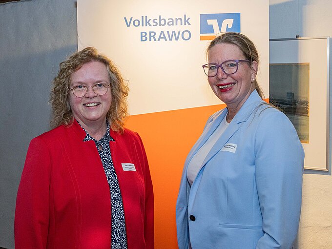 Auf dem Foto sieht man zwei Frauen, die in die Kamera lächeln. Die Frau links hat blonde lockige Haare, trägt eine Brille und ein rotes Jacket mit einer gemusterten blauen Bluse. Die Frau rechts daneben hat blonde Haare und trägt einen hellblauen Blazer mit einem weißen Oberteil.