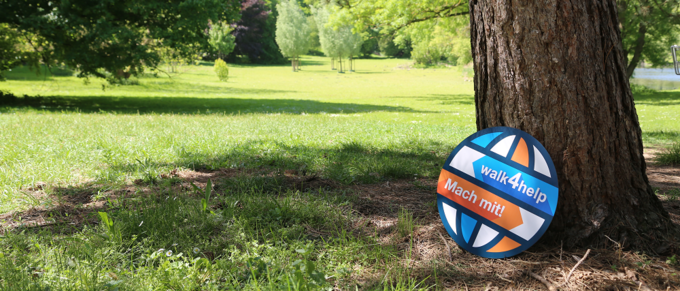 Eine blau-orangene Pappscheibe mit der Aufschrift "walk4help" lehnt an einem Baumstamm in einem Park. Die Rasenfläche dahinter ist kräftig grün und sonnenbeschienen.