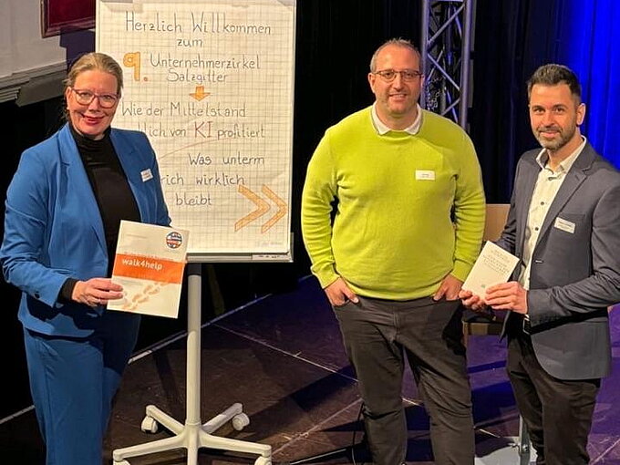 Das Bild zeigt drei Personen, die neben einem Flipchart stehen, das die Begrüßung zu dem Unternehmerzirkel Salzgitter zeigt. Die Frau links trägt einen blauen Hosenanzug und hält eine Broschüre in der Hand. Der Mann in der Mitte trägt einen grünen Pullover, während der Mann rechts einen dunklen Anzug trägt. Alle drei blicken freundlich in die Kamera.