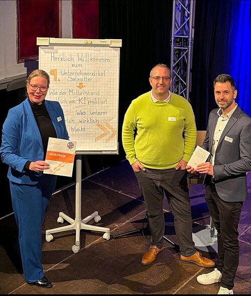 Das Bild zeigt drei Personen, die neben einem Flipchart stehen, das die Begrüßung zu dem Unternehmerzirkel Salzgitter zeigt. Die Frau links trägt einen blauen Hosenanzug und hält eine Broschüre in der Hand. Der Mann in der Mitte trägt einen grünen Pullover, während der Mann rechts einen dunklen Anzug trägt. Alle drei blicken freundlich in die Kamera.