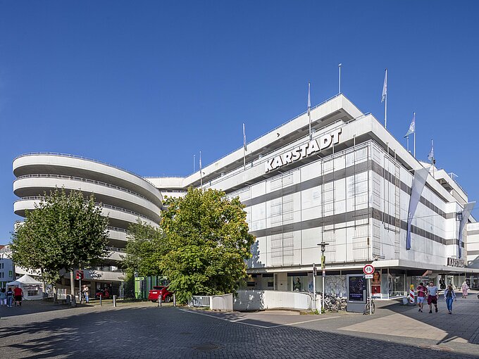 Großes Kaufhaus unter blauem Himmel mit belebter Straße im Vordergrund.