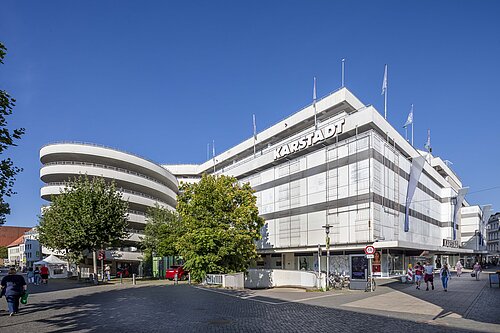 Großes Kaufhaus unter blauem Himmel mit belebter Straße im Vordergrund.