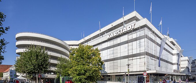 Großes Kaufhaus unter blauem Himmel mit belebter Straße im Vordergrund.
