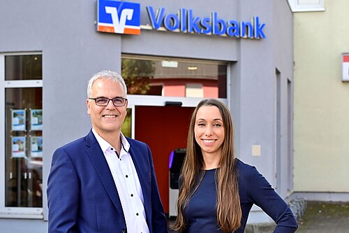 Ein Mann und eine Frau stehen lächelnd vor einem Gebäude mit einem "Volksbank" Zeichen. Der Mann trägt einen blauen Anzug und eine Brille, die Frau ein dunkles Kleid. Im Hintergrund ist ein Eingang und Schaufenster mit Werbeplakaten zu sehen.
