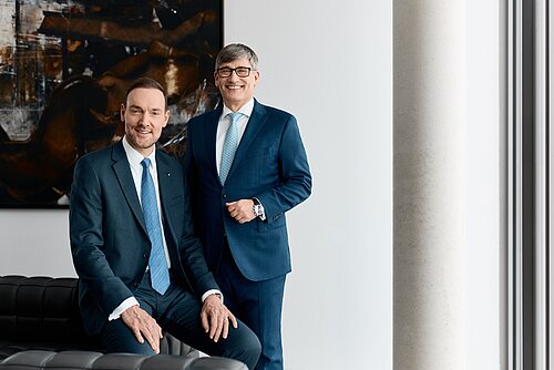 Zwei Männer in Anzügen posieren lächelnd in einem modernen Büro vor einem abstrakten Gemälde.