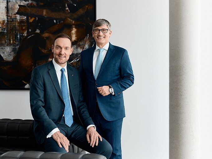 Zwei Männer in Anzügen posieren lächelnd in einem modernen Büro vor einem abstrakten Gemälde.