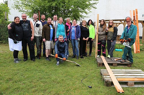 Eine Gruppe von fünfzehn Personen posiert lächelnd auf einer Wiese. Einige tragen Arbeitskleidung, darunter helle Jacken und Handschuhe. Eine Person kniet vorn auf dem Boden mit einer Schaufel. Rechts steht ein Mann mit einer Motorsäge auf Paletten. Im Hintergrund sind Bäume und eine Wand zu sehen. Holzbretter und Werkzeuge liegen verstreut auf dem Boden.