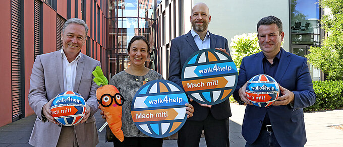 Auf demBild sieht man drei Männer und eine Frau, die in die Kamera schauen und verschiedene Gegenstände in den Händen halten. Der Mann ganz links trägt ein graues Jacket und hält einen orange-blauen Ball. Die Frau rechts daneben trägt eine Bluse und hält eine Plüschkarotte mit Brille sowie ein rundes orange-blaues Schild. Der Mann rechts neben ihr trägt ein ähnliches Schild und ist in einem dunkelblauen Anzug gekleidet. Der Mann ganz rechts trägt ebenfalls einen dunkelblauen Anzug und hält einen orange-blauen Ball in den Händen.