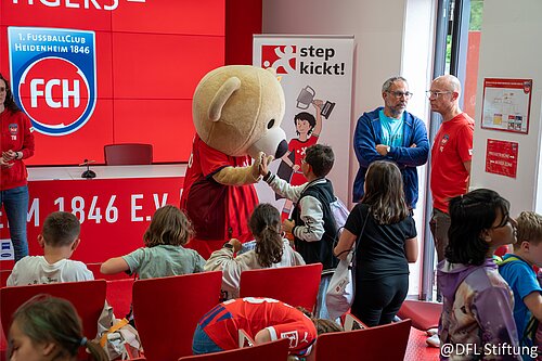 Maskottchen begrüßt Kinder in einem Raum mit FCH-Logo, während zwei Erwachsene im Hintergrund stehen.