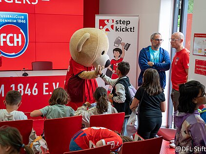 Maskottchen begrüßt Kinder in einem Raum mit FCH-Logo, während zwei Erwachsene im Hintergrund stehen.