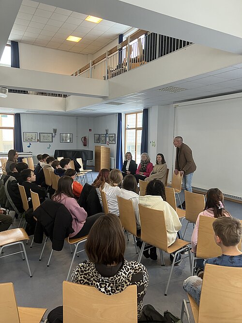 Schüler sitzen in einem Raum und hören mehreren Rednern zu, die vorne sprechen.