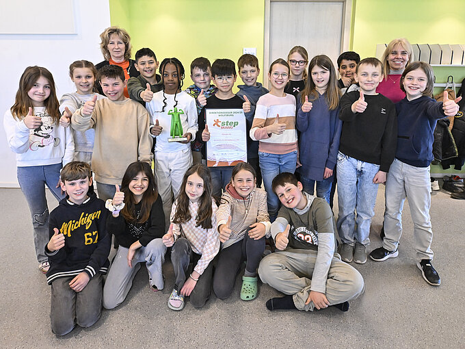 Auf dem Bild sieht man eine Gruppe von Kindern, die in die Kamera lächeln. Einige von Ihnen zeigen mit dem Daumen nach oben und zwei Kinder halten jeweils den step BRAWO-Pokal und die Urkunde in der Hand. Hinter den Kindern steht links eine in orange gekleidete blonde Frau und rechts eine in pink gekleidete blonde Frau.