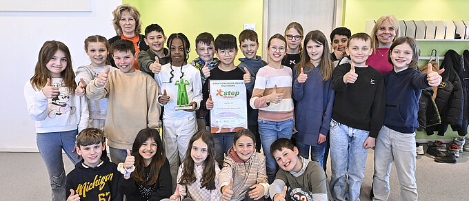 Auf dem Bild sieht man eine Gruppe von Kindern, die in die Kamera lächeln. Einige von Ihnen zeigen mit dem Daumen nach oben und zwei Kinder halten jeweils den step BRAWO-Pokal und die Urkunde in der Hand. Hinter den Kindern steht links eine in orange gekleidete blonde Frau und rechts eine in pink gekleidete blonde Frau. 