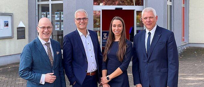 Vier Personen in formeller Kleidung stehen vor einem Gebäude mit dem Logo und Schriftzug der Volksbank. Das Gebäude hat große Fenster und einen Eingang. Die Personen lächeln in die Kamera.