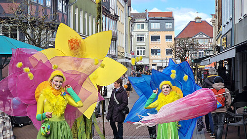 Straßenkünstler in bunten Blumen- und Blütenkostümen bei einem Stadtfest spazieren durch eine belebte Straße.