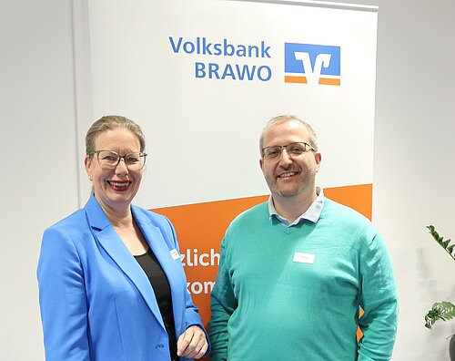 Zwei Personen lächeln vor einem Volksbank BRAWO Banner.