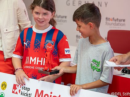 Zwei Kinder halten eine rote Fußball-Trophäe und lächeln in die Kamera bei einer Veranstaltung.
